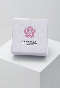 White textured jewelry box with a pink floral logo and black text "SENSORA JEWELRY" on the front, resting on a white pedestal.