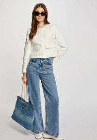Giacca cropped bianca e testurizzata con due tasche frontali, abbinata a jeans a vita alta blu con gamba svasata. In mostra anche una borsa a tracolla blu trapuntata.