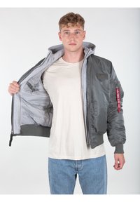 Giacca bomber grigia con cappuccio e foderatura grigia, polsini elasticizzati, due tasche laterali e un'etichetta rossa sulla manica. Indossata aperta sopra una camicia beige.
