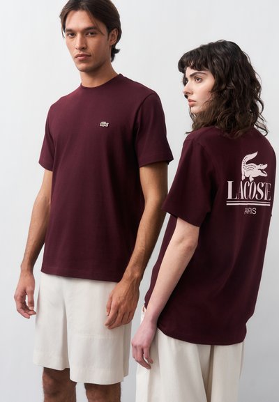 Два модела носят еднакви бордо Lacoste тениски и бели къси панталони; предната тениска показва малко лого, а задната тениска има голям надпис Lacoste Paris.