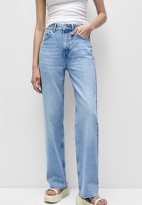 Persona vestida con jeans rectos de color azul claro, top de tirantes blanco y sandalias de plataforma crema, con una mano en el bolsillo, frente a un fondo plano.