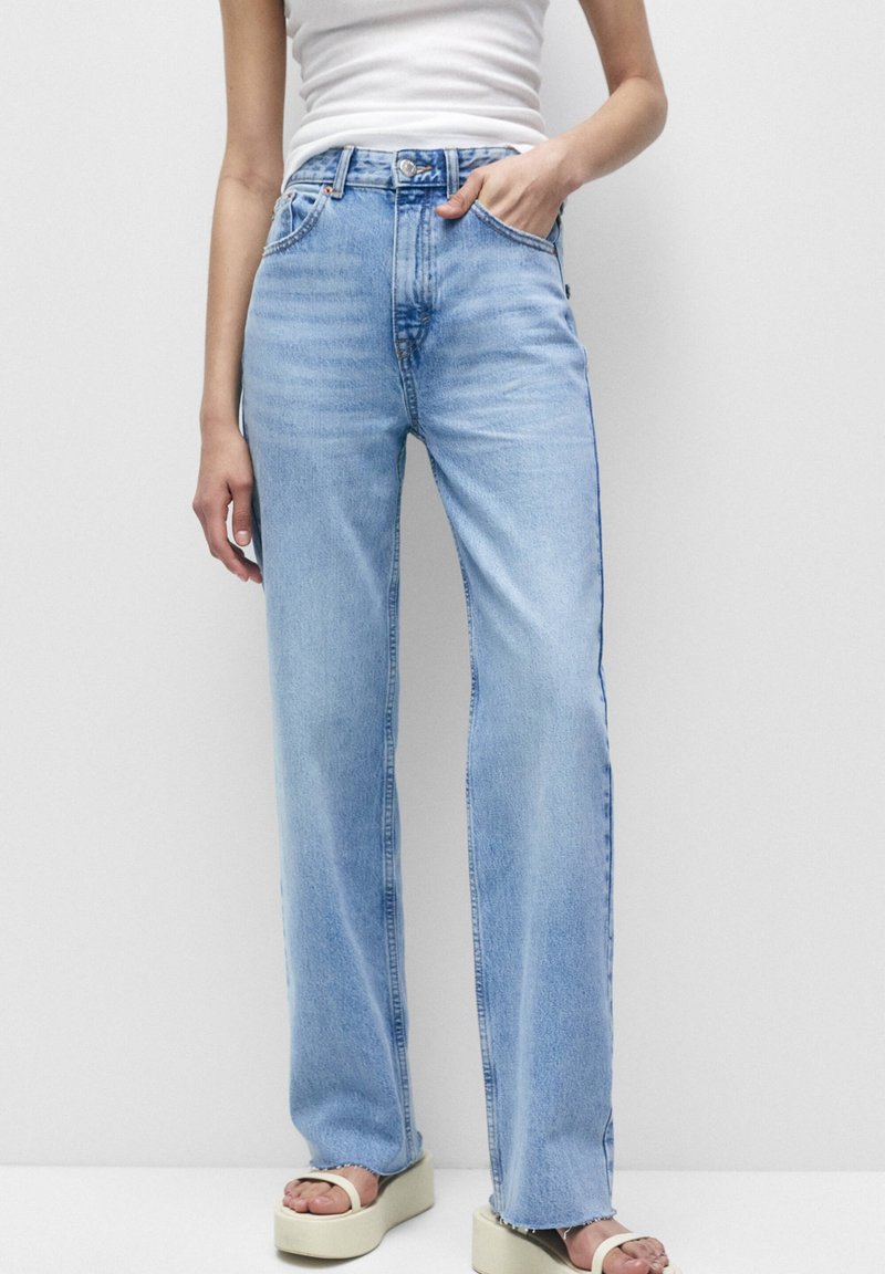 Persona vestida con jeans rectos de color azul claro, top de tirantes blanco y sandalias de plataforma crema, con una mano en el bolsillo, frente a un fondo plano.