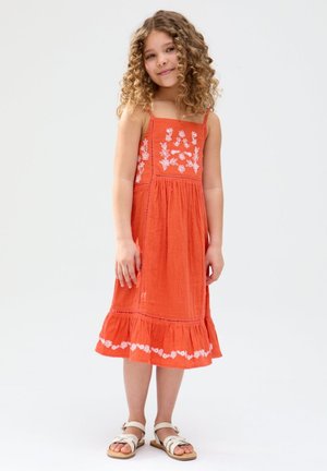 Junges Mädchen mit lockigem Haar, das ein oranges ärmelloses Kleid mit weißer Blumenausfertigung und weiße Sandalen trägt, steht vor einem einfarbigen Hintergrund.