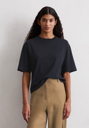 T-shirt nera in cotone con maniche corte, scollatura rotonda e vestibilità rilassata, abbinata a pantaloni beige chiaro in tessuto intrecciato.