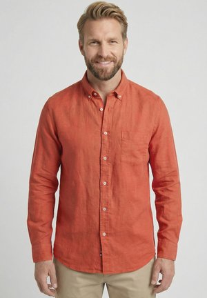 Lächelnder Mann mit kurzem blondem Haar und Bart, der ein orangefarbenes langärmeliges Hemd und beige Hosen vor einem schlichten Hintergrund trägt.