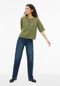 Zielony kardigan z mieszanki wełny z krótkimi rękawami, zapinany na pięć guzików z przodu. W zestawie z niebieskimi dżinsami o prostych nogawkach i czarnymi butami.