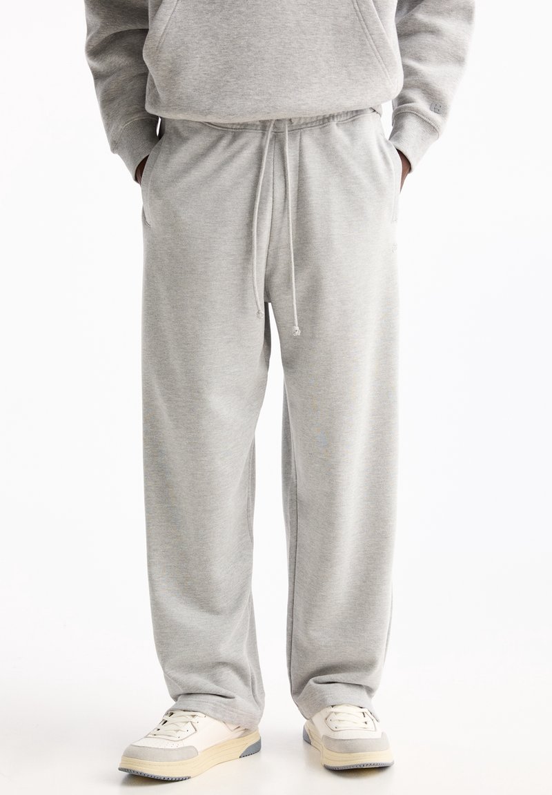 Personne portant un pantalon de survêtement gris clair et un sweat assorti, les mains dans les poches, debout en baskets blanches sur fond blanc.