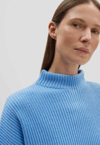 Hellblaues Ripp-Strickoberteil mit hohem Kragen, das ein strukturiertes Strickmuster aufweist. Der Stoff wirkt weich und eng anliegend am Ausschnitt.