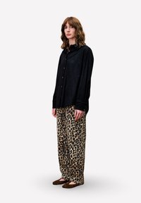 Frau steht da und trägt ein lockeres schwarzes Button-Down-Hemd, Leopard-Print-Hosen im weiten Schnitt und braune flache Schuhe vor einem schlichten Hintergrund.