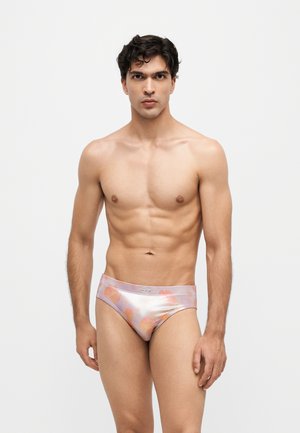 Joven con cabello corto y oscuro que lleva bragas brillantes de color rosa claro y naranja con estampado, de pie frente a un fondo blanco liso.