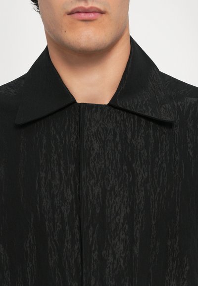 Schwarze texturierte Jacke mit spitzem Kragen und einem Vorderreißverschluss. Der Stoff weist ein dezentes, variierendes Muster auf, das ein zerknittertes Aussehen verleiht.