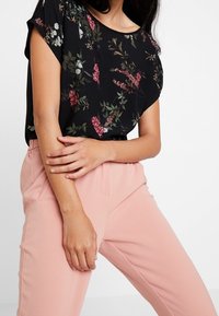 Černá blůza s květinovým vzorem, která obsahuje růžové a bílé květy, spárovaná s jemně růžovými zúženými kalhotami, které mají uvolněný střih a kapsy.