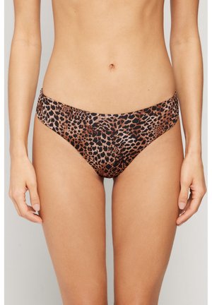 Mujer con braguitas de bikini con estampado de leopardo, de pie con los brazos relajados a los lados contra un fondo liso.