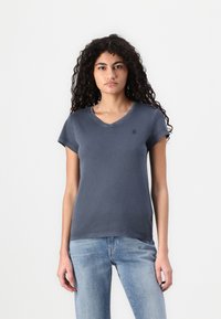 T-shirt de algodão cinza com decote em V e mangas curtas, apresentando um pequeno logotipo no peito e uma bainha reta. Combinado com jeans azul claro.