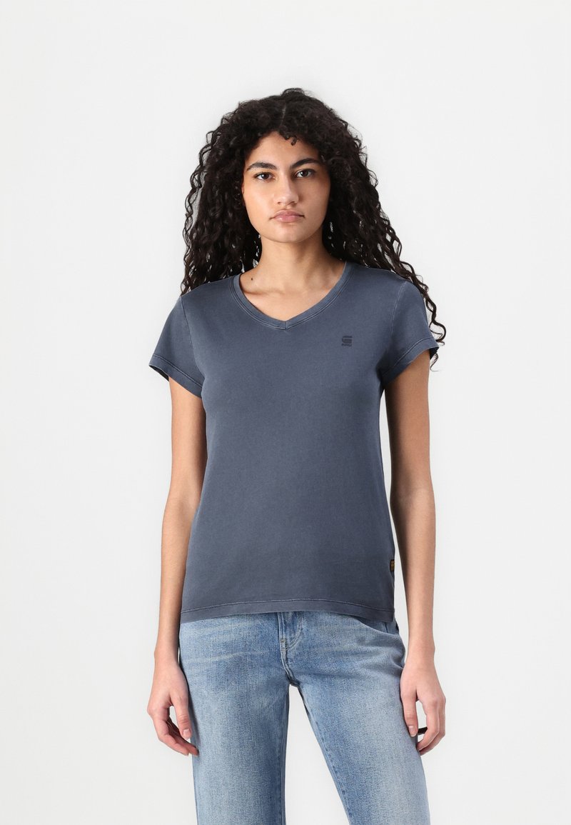 T-shirt de algodão cinza com decote em V e mangas curtas, apresentando um pequeno logotipo no peito e uma bainha reta. Combinado com jeans azul claro.