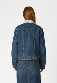 Τζάκετ από τζιν με ελαφριά μπλε γιακά από sherpa, σκούρο πλύσιμο, κλασική γραμμή, χάλκινα κουμπιά και πίσω τσέπες με κόκκινη λεπτομέρεια.