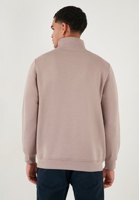 Sudadera marrón claro con un cuello alto, mangas largas y puños acanalados. Textura suave, corte informal y un color sólido sin patrones.