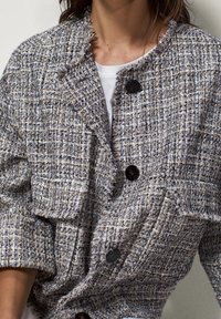 Tkany żakiet z tweedu w kolorze beżowym i niebieskim, z teksturowanym wykończeniem, dwoma dużymi kieszeniami naszytymi i czarnymi guzikami. Krawędzie są postrzępione.