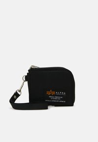 Portefeuille en tissu noir avec une fermeture zippée, doté d'une poignée de poignet et d'un patch logo orange avec du texte sur le devant. Forme rectangulaire compacte.