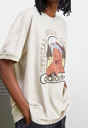 T-shirt beige oversize avec un motif représentant un ours, des arbres, des montagnes, le soleil, des oiseaux, ainsi que le texte "Official Guide" et le logo "adidas", porté par une personne les mains dans les poches.