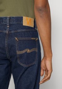 Tmavě modré džíny z denimu s oranžovým prošitím. Na pasu je hnědá kožená záplata a na zadním kapse dekorativní prošití.