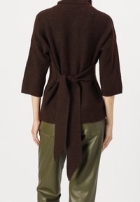 Pull tricot marron texturé avec une ceinture à la taille et des manches ¾, porté sur un pantalon vert brillant. Vue de dos montrant le détail de la ceinture.