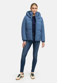 Lichtblauwe donsjacket met een capuchon, voorzien van een gewatteerd ontwerp, voorkzakken en een relaxte pasvorm, gecombineerd met donkere denim en sneakers.