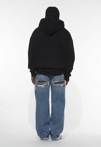 Black oversized hoodie with a large hood, paired with distressed light blue jeans featuring rips and patches. White sneakers visible.