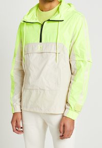 Kurtka pulower w neonowej zieleni i beżu, z przednią kieszenią, regulowanym kapturem i elastycznymi mankietami. Gładki, lekki materiał, sportowy krój.