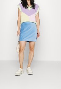 Blauwe corduroy mini-rok met een zijsplit, gecombineerd met een meerkleurige gebreide vest met geometrische patronen in lavendel en groen. Witte platform schoenen.