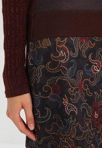 Un pull bordeaux tricoté avec une texture côtelée, assorti à une jupe à motifs avec des motifs floraux en rouge, bleu et or sur un fond noir.