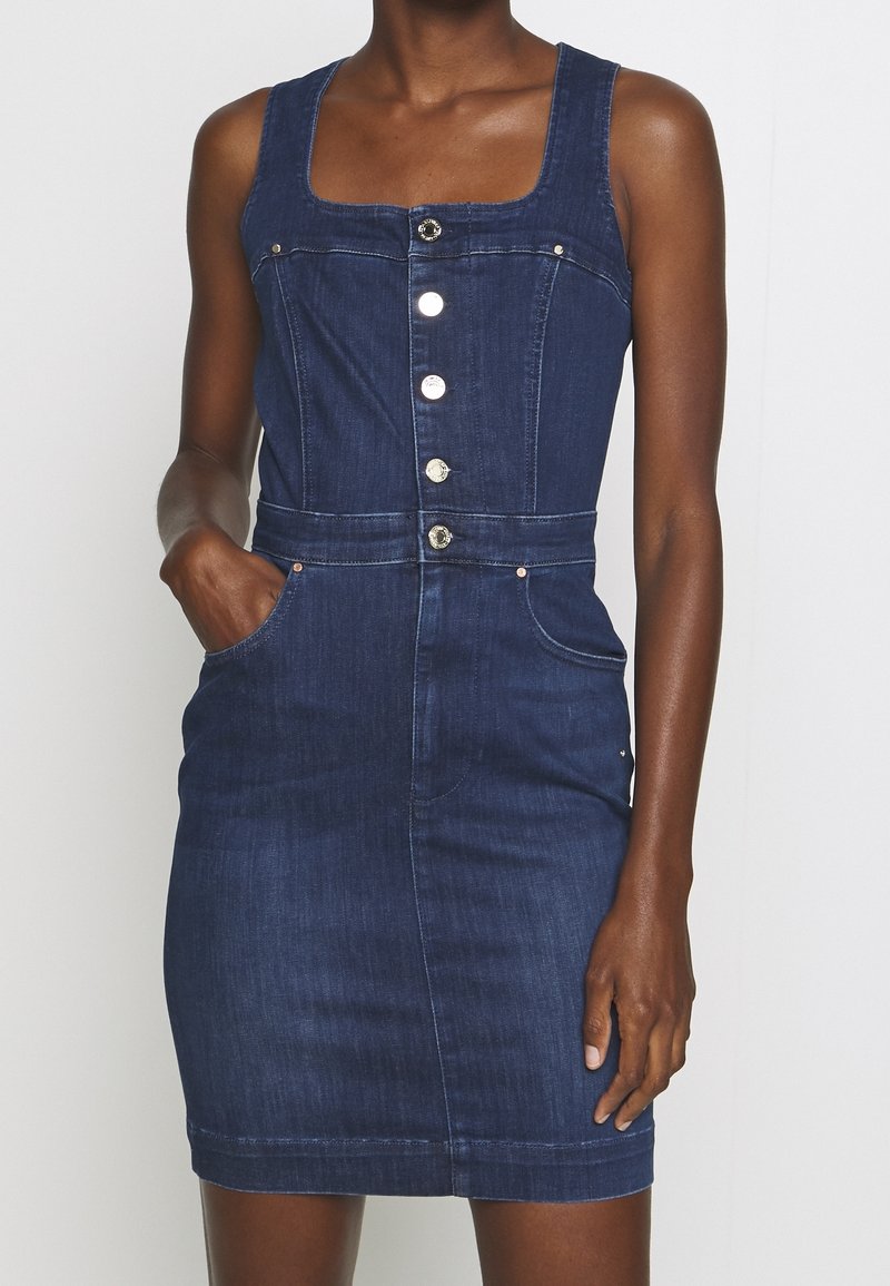 Mörk denim, ärmlös klänning med fyrkantig halsringning, försedda med framknappar, fickor och en figurnära silhuett. Slät textur och knälång.