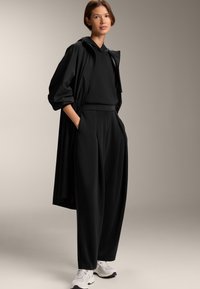 Fekete kapucnis dzseki laza ujjakkal, párosítva egy azonos fekete pulóverrel és magasan ültetett, pliszézett nadrággal. Fehér sportcipők teszik teljessé a megjelenést.