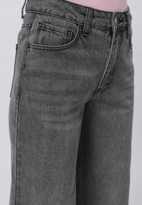 Graue, hoch taillierte Jeans, die mit einem rosa, eingesteckten Oberteil getragen werden, mit Nahaufnahme der vorderen Taschen, des Taillenbunds und des Knopfverschlusses.