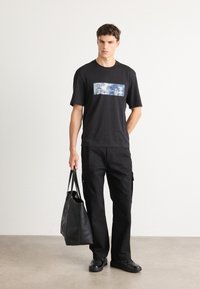 Ung mand iført sort grafisk t-shirt og sorte cargo bukser, som holder en stor sort læder tote bag, stående mod en ensfarvet baggrund.