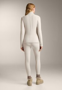 Witte, nauwsluitende langemouwtop met een hoge kraag, gecombineerd met bijpassende legging; het model draagt ook tan enkellaarsjes met bontaccenten.