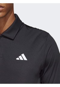 Černé polo tričko s límečkem, vyrobené z hladké látky. Na levé straně je bílý trojúhelníkový logo.