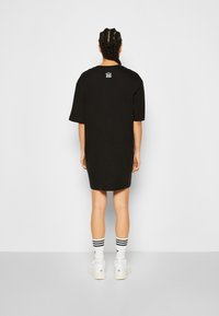 Black oversized t-shirt dress with short sleeves, featuring a small white logo on the back. The fabric appears smooth, suitable for casual wear.