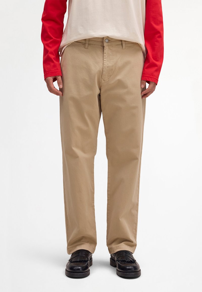 Άτομο που φοράει μπεζ παντελόνι ίσιας γραμμής, μαύρα μοκασίνια και κρεμ πουκάμισο με μακριά κόκκινα μανίκια, στέκεται μπροστά σε λευκό φόντο.