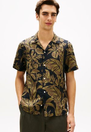 Junger Mann trägt ein schwarzes kurzärmliges Hemd mit großen olivgrünen tropischen Blättern und Blumenmuster.