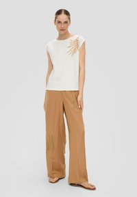 Haut blanc à manches courtes avec un motif de feuille brodé en or sur le côté gauche, associé à un pantalon large couleur brun clair présentant une broderie similaire.