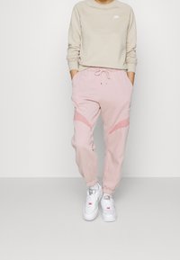 Osoba oblečená v béžové mikině Nike, světle růžových trenbirkách Nike se stahovací šňůrkou a bílých teniskách Nike, stojí s rukama v kapsách.