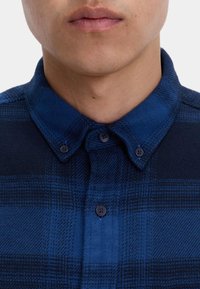 Zbliżenie na osobę ubraną w niebieską i granatową koszulę w kratę z zapiętą kołnierzykiem.