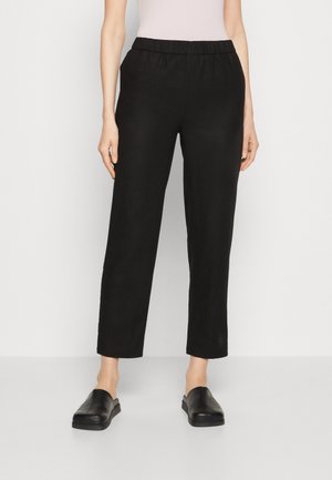 Onderlichaam van een persoon die zwarte rechte broek met elastische taille en zwarte instapschoenen draagt, staand tegen een effen lichte achtergrond.
