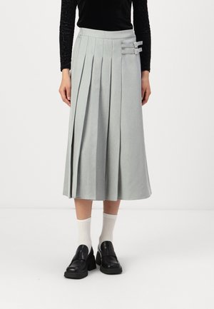 Femme portant une jupe midi plissée gris clair avec deux boucles sur les côtés, des chaussettes blanches, des mocassins noirs à talons et un haut noir à manches longues.