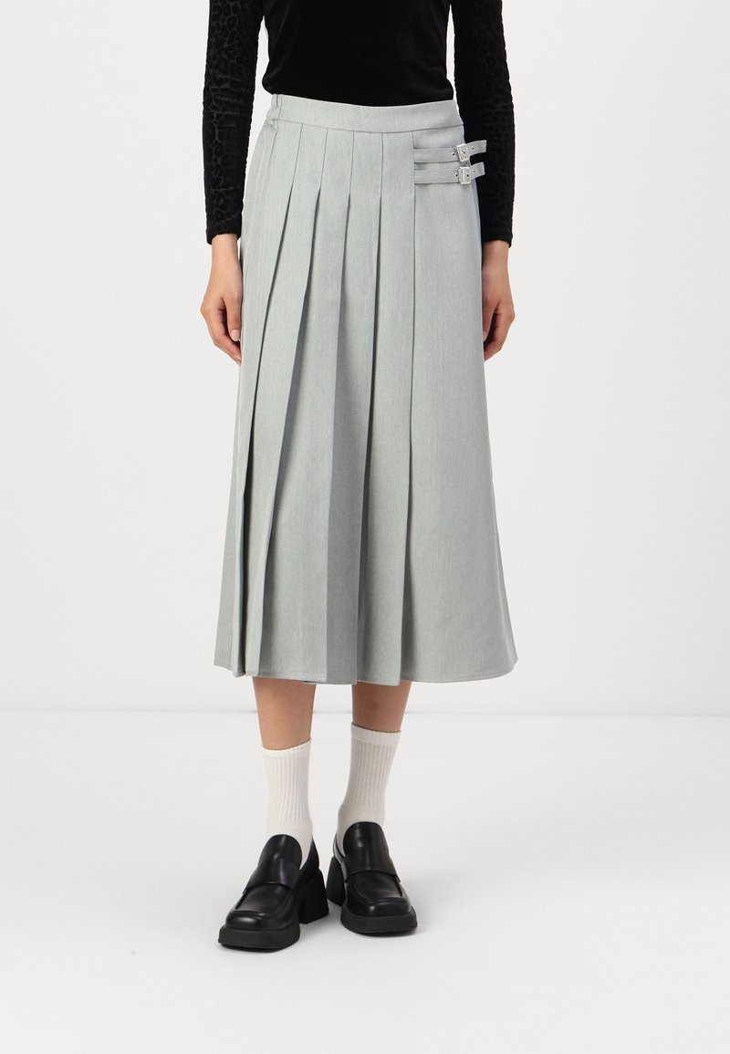 Femme portant une jupe midi plissée gris clair avec deux boucles sur les côtés, des chaussettes blanches, des mocassins noirs à talons et un haut noir à manches longues.