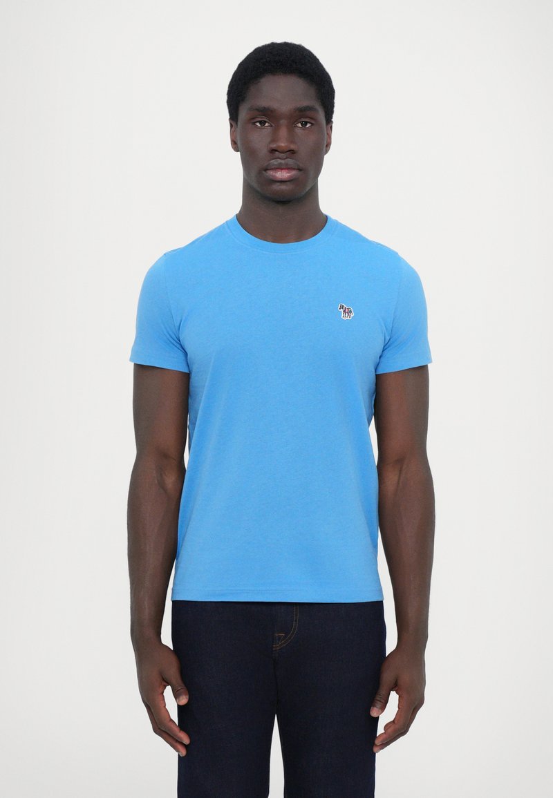 Tricou scurt cu mâneci, de culoare albastră, confecționat din bumbac moale, având un mic logo brodat pe piept, asortat cu blugi negri.