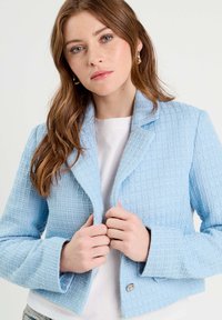 Veste texturée bleu clair au design court, avec un col cranté et une fermeture à boutons. Le mannequin tient les revers tout en portant un top blanc en dessous.