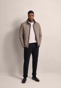 Beige Steppjacke mit Reißverschluss, hohem Kragen und gerippter Textur, kombiniert mit schwarzen Hosen und schwarzen Slippern.