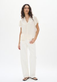 Blusa beige chiaro a maniche corte con scollo a V e maniche arricciate, abbinata a pantaloni bianchi a gamba larga e infradito neri.